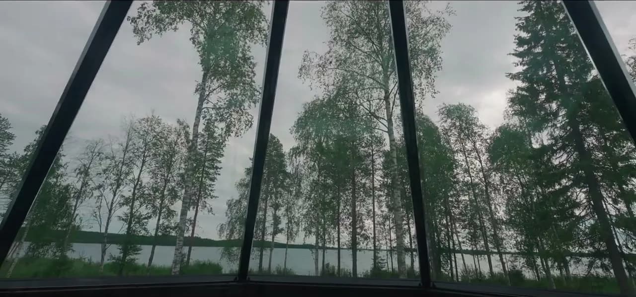 A glass cabin looking out over birch trees and a Lapland lake under bright northern sky