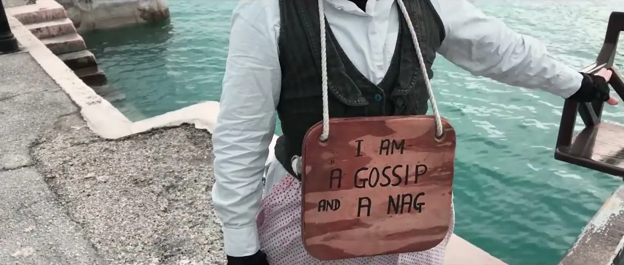 A reenactor holding a sign reading 'I am a gossip and a nag' at the St George's ducking stool