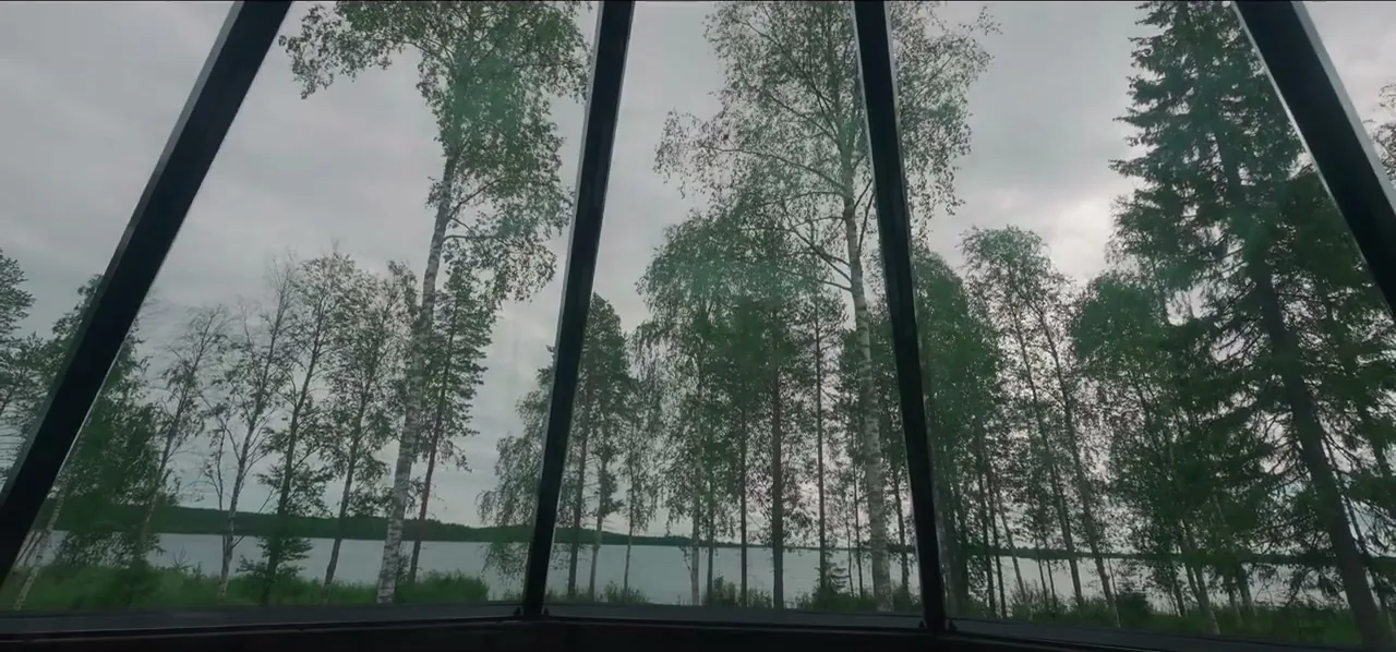 A glass cabin looking out over birch trees and a Lapland lake under bright northern sky